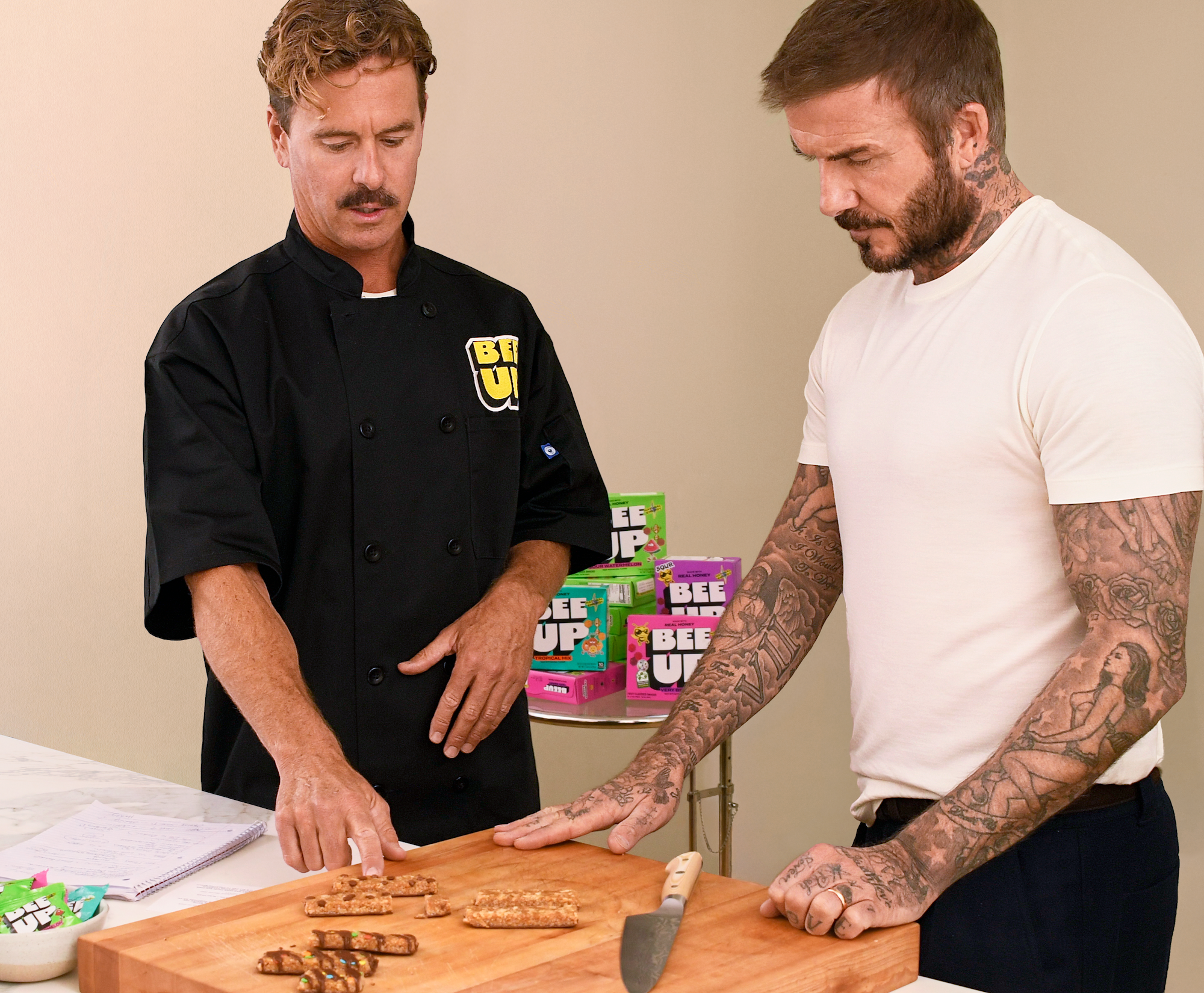 Chef in black BEEUP uniform and David Beckham examining energy bars on wooden cutting board, with stacked BEEUP product boxes in background.