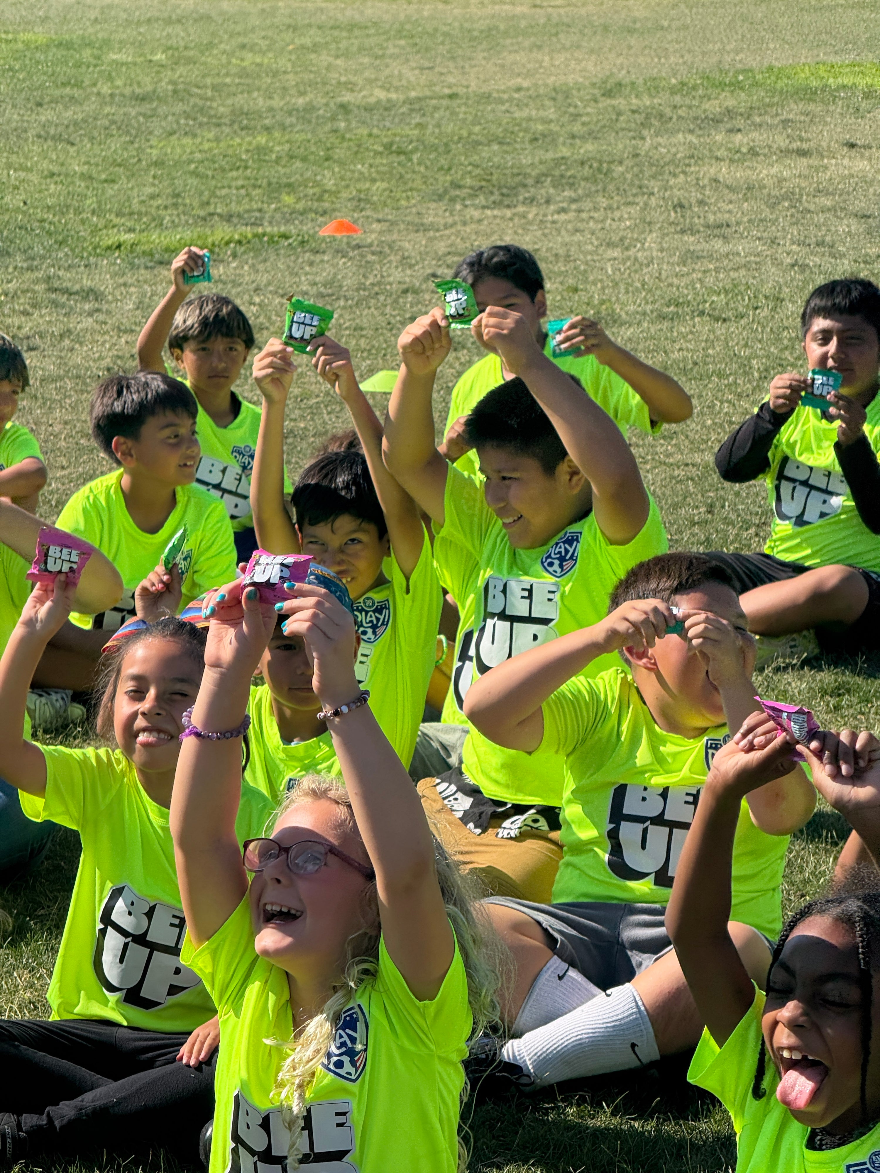 Excited children in BEEUP soccer uniforms, holding up BEEUP honey fruit snacks, while sitting in a soccer field