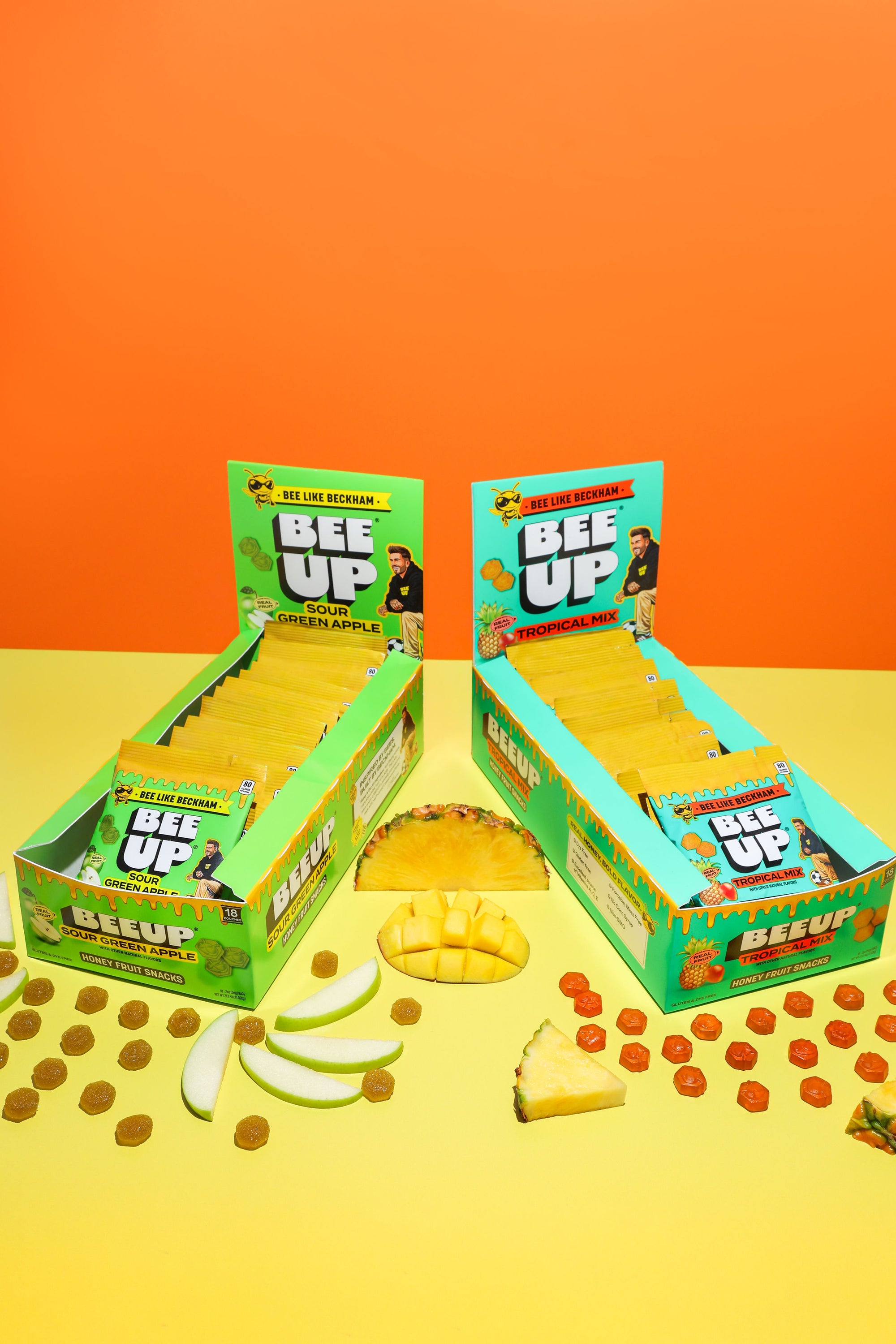 Two boxes of 'BEE UP' caddy's with Sour Green Apple on the left side and Tropical Mix being on the right side on a yellow and orange gradient background with fruits surrounding the two boxes.