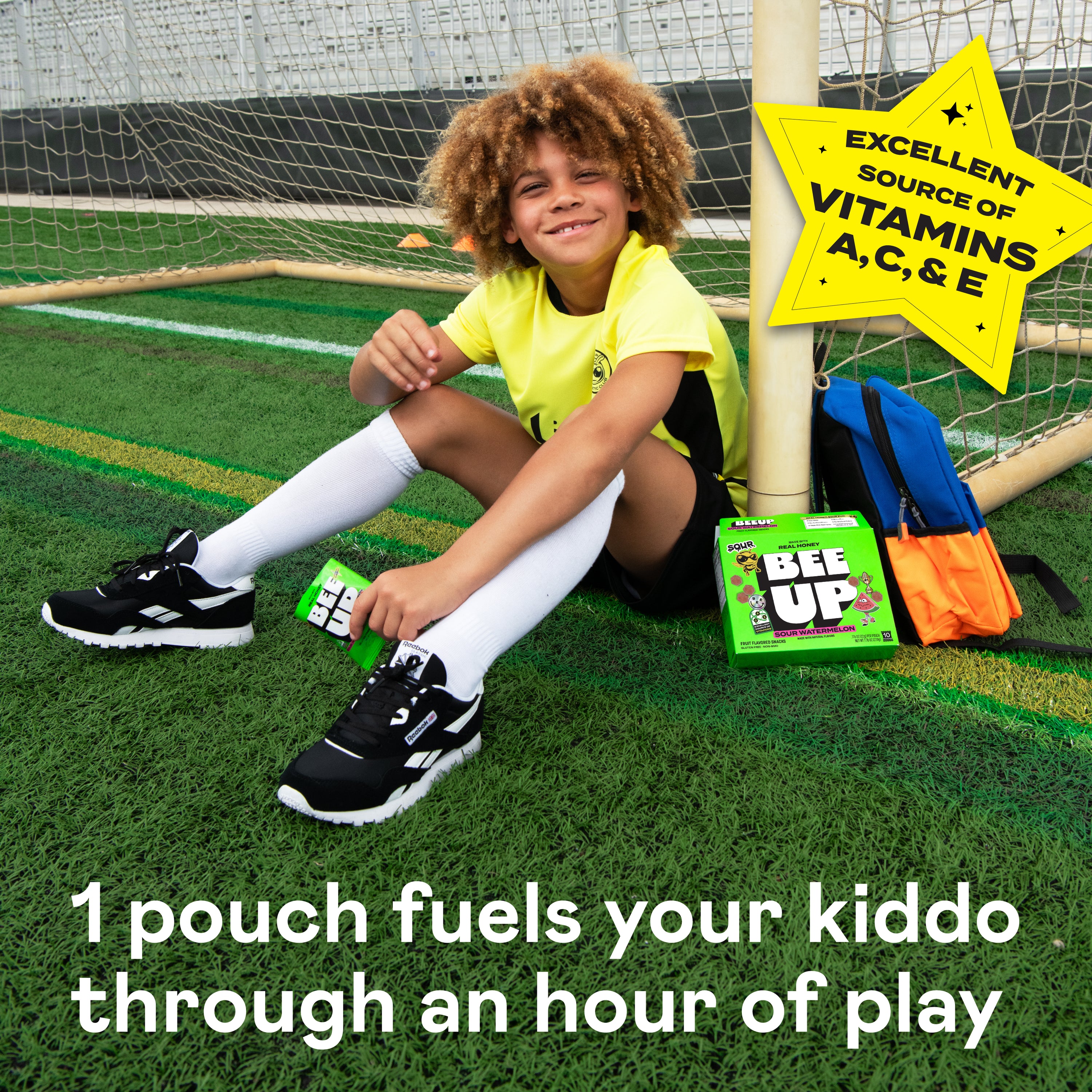 Child is sitting on a soccer field by a goal post, enjoying sour berry BEEUP honey fruit snacks. Text at the bottom says, "1 pouch fuels your kiddo through an hour of play" There is a star shaped sticker at the top right stating, "Excellent source of vitamins A, C, & E"